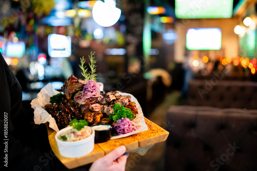 Tableau sur toile A person holds a wooden platter piled high with a variety of grilled meats, salads, and sides in a dimly lit restaurant or bar setting