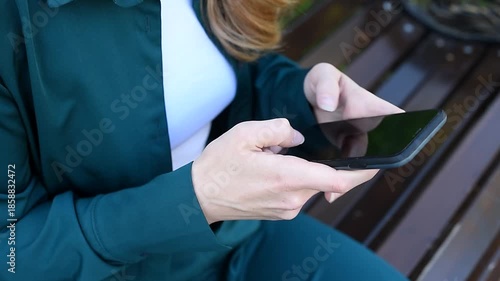 young woman holds smartphone in hands, texting, watching content, sitting on bench in park, close-up view, no face, copy space