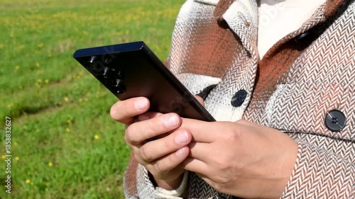 young woman holds smartphone in hands, standing on country road in green field in sunny summer day, beige plaid shirt, travel blogger, close-up view, no face, copy space