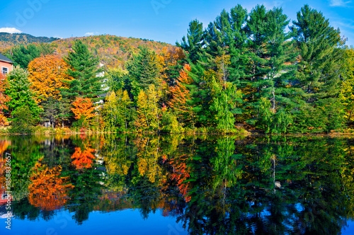 Scenic autumn view of colorful trees reflecting on a calm lake at Mont-Tremblant, Quebec. Vibrant fall foliage, blue sky, and serene nature create a peaceful landscape.