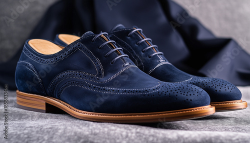 Pair of dark blue suede brogue shoes with leather laces rests on grey tweed fabric. These classic formal footwears have elegant wingtip