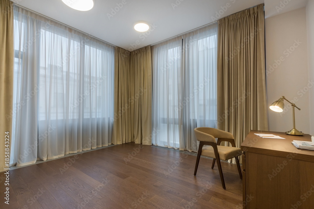 Fototapeta premium Bright room has large windows covered with light curtains. A wooden desk and a chair are placed on the wooden floor. The room appears empty and open, suitable for various activities