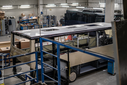 Industrial vehicle manufacturing workshop with metal bus frames and body structures under assembly. Large factory interior shows fabrication, welding, and custom vehicle production in progress.