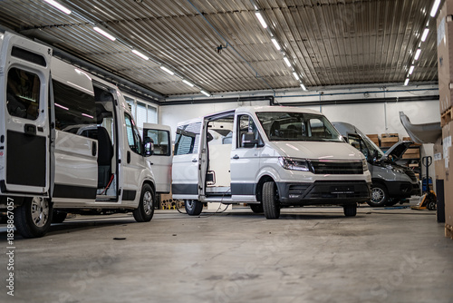 Commercial vans in industrial workshop during conversion and assembly. Vehicle interiors exposed, doors open, showing custom transport production, maintenance, and manufacturing process.
