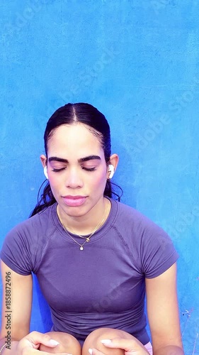 Tired female athlete sitting against a blue wall listening to music and feeling stressed