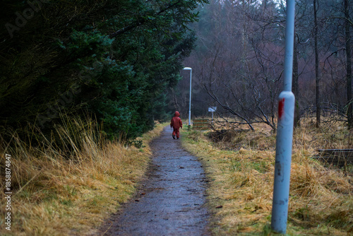 Child in a bright red raincoat walking away along a path in a gloomy misty autumn forest