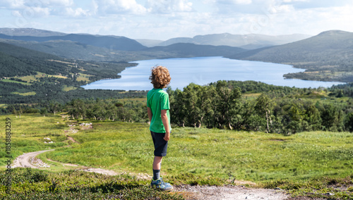 A boy with curly blond hair looks out from a hiking trail on a mountainside. He sees a lake surrounded by mountains in Trollheimen, Norway.
