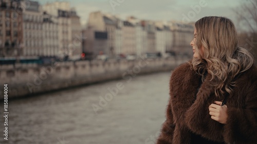 A woman wearing a brown fur coat stands overlooking the Seine River in Paris France with a cityscape background. This image is suitable for travel tourism and romantic getaway themes.
