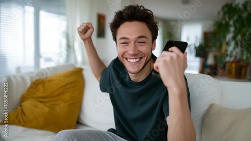 young man sits on a soft white sofa in a bright, cozy living room, looking at his phone with an expression of pure excitement as he raises one hand in a triumphant fist. He wear
