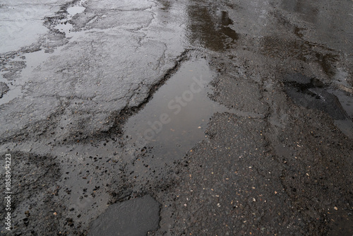 Fotografie Close-up view of a severely damaged asphalt road covered in cracks, deep potholes filled with rainwater, and scattered gravel, showcasing poor road maintenance on a gloomy, overcast day