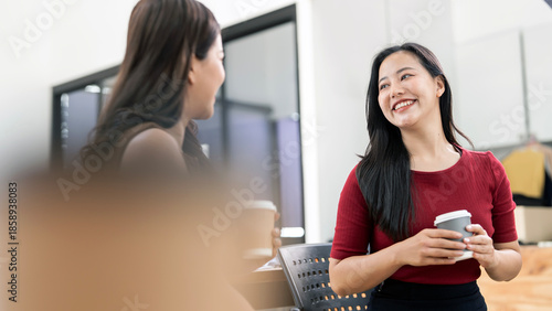 Two smiling Asian businesswomen having a friendly conversation and holding coffee cups in a modern office workspace.