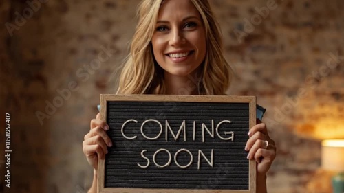 A joyful, expectant young woman with radiant blonde hair smiles brightly directly at the camera while holding a personalized letter board displaying the hopeful message 