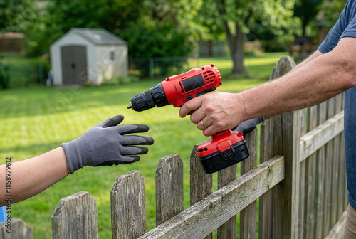 Close up male neighbor hands passing red cordless power drill to person in work glove over wooden fence, concept of lending tools, diy home improvement and helpful community sharing equipment.