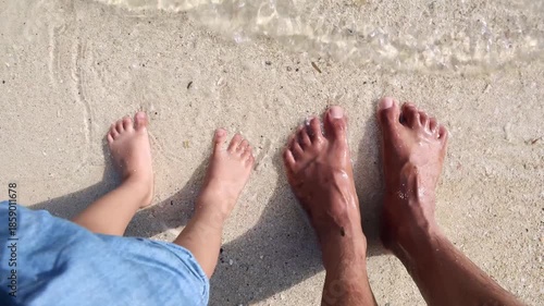 father and child feet in shallow sea water on sandy beach top view