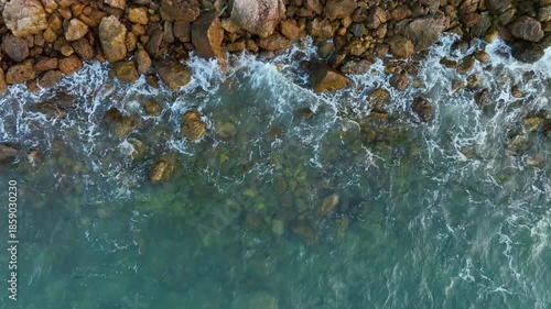 Angle drone shot capturing the beautiful contrast between the deep blue sea foam and the brown textured boulders along the shoreline, creating a natural abstract pattern.concept nature