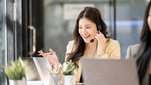 Tableau sur toile Professional Asian woman with headset working in call center, friendly customer service agent talking to client via video call, remote consultant in modern office