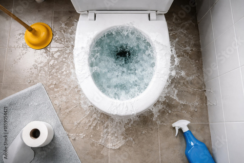 Conceptual home maintenance photo of clogged toilet emergency. Water spilling from overflowing bowl onto floor creates messy, dramatic composition from top down perspective