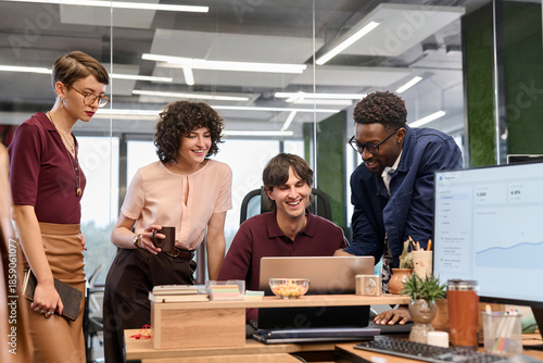 Diverse group of young adult men and women collaborating around laptop in modern office, smiling and discussing project while standing and sitting together, computer monitor visible in foreground