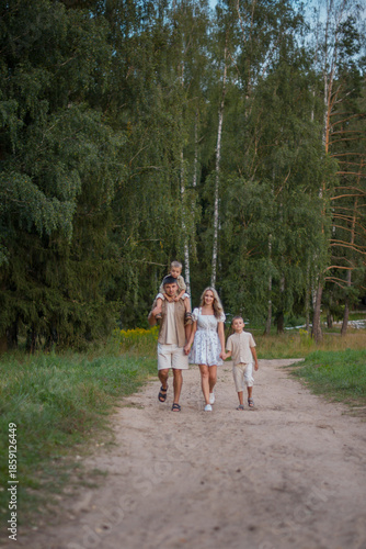 A family walks along a dirt path in a forest. Two children, a boy and a girl, are accompanied by their parents. The scene is serene and natural.