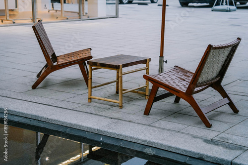 Outdoor relaxation area on concrete road by water decorated with aged wooden chairs and small table.