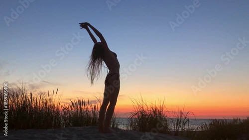 Silhouette of woman stretching at sunset on beach with grasses in foreground. Calm fitness moment.