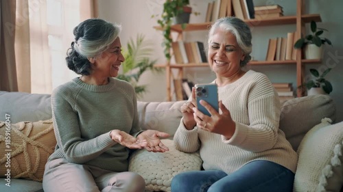 Senior women laughing together while using a smartphone on a cozy sofa at home