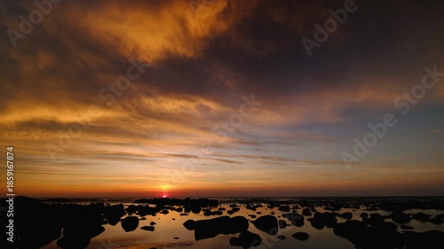 Peaceful time-lapse style footage of sun slowly setting behind sea horizon, coloring dramatic clouds in orange and gold and reflecting in calm water with dark rocks in foreground