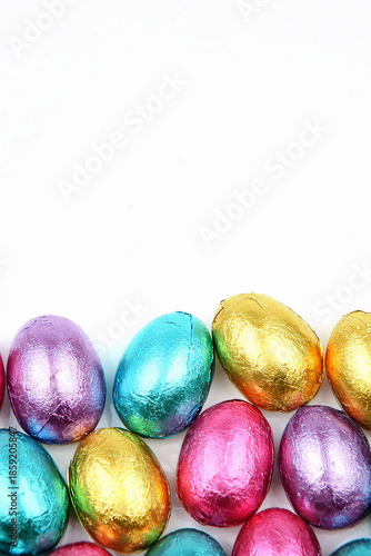 Pile of foil wrapped chocolate easter eggs in pink, blue & lime green with a white background.