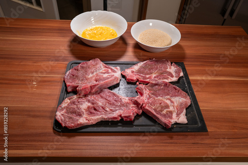 Overhead food photography showing the preparation of breaded meat with raw pork steaks on a black cutting board, a bowl of breadcrumbs, and a bowl of beaten eggs 