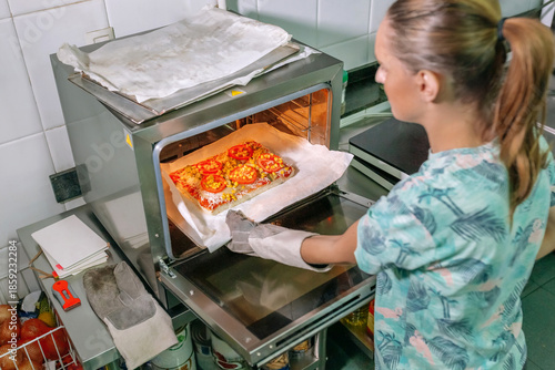 Unrecognizable female chef skillfully removing a freshly baked pizza from professional oven in commercial kitchen, expertly preparing a delicious meal for eager customers