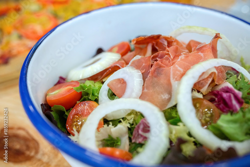 Detail of prosciutto salad mixing fresh green lettuce, cherry tomatoes, and onion rings in a white enamel bowl, on natural wood table in vegan restaurant. Organic and healthy fast food concept.