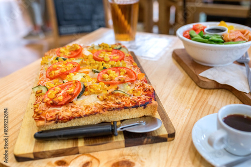 Rectangular vegetarian pizza with melted cheese, tomato, zucchini, and corn kernels, being served with a pizza cutter, a fresh poke bowl, and a coffee on a wooden table inside of italian restaurant