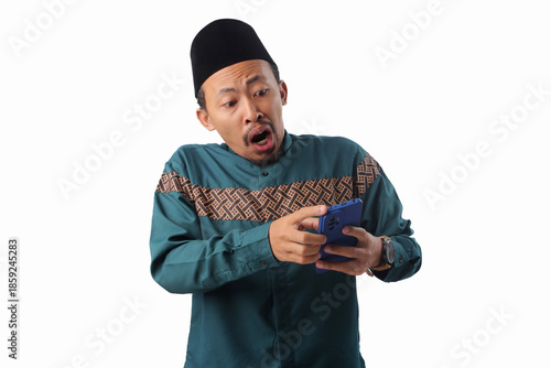 Frustrated Asian Muslim man in green traditional outfit looking stressed after receiving unexpected news or notification on his phone. Isolated on white background.