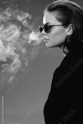 Black and white portrait of a young woman wearing sunglasses exhaling smoke in a dark background.