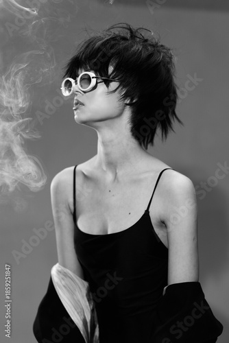Black and white portrait of a stylish woman with short hair wearing round sunglasses and a black dress with smoke in the background.