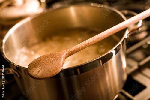 A wooden cooking spoon lies on a stainless steel pot in which water is boiling on a black gas stove.