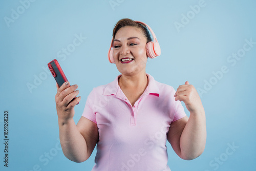 portrait of adult latin woman listening to music and using headphones while holding mobile phone on blue background in Mexico Latin America, hispanic and mexican female