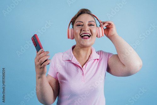 portrait of adult latin woman listening to music and using headphones while holding mobile phone on blue background in Mexico Latin America, hispanic and mexican female