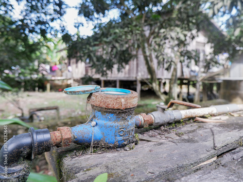 ​Close-up of a residential water meter with a blue lid and valve on wooden background. Concept of water consumption, utility management, and plumbing system in a home garden
