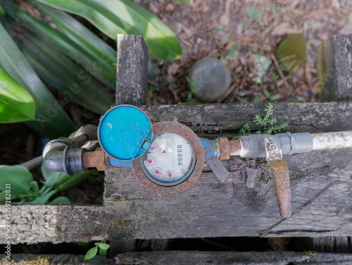 ​Close-up of a residential water meter with a blue lid and valve on wooden background. Concept of water consumption, utility management, and plumbing system in a home garden