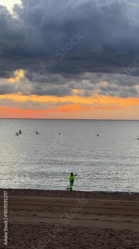 Sunset beach landscape with dramatic storm clouds over calm sea, workers cleaning sand in the foreground and paddle surfers on the horizon, environmental care concept, coastal nature scene and cinemat