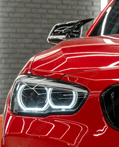 Front BI-LED headlight on a red sport car. Gloss red premium car LED headlights close-up. Modern sport car headlight. BI-LED headlights of a modern and expensive red car, hood and radiator grille. 