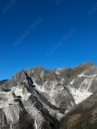 Carrara marble quarry in the Apuan Alps, Italy, dramatic mountain landscape with white stone cuts, rugged cliffs and deep valleys under a clear blue sky
