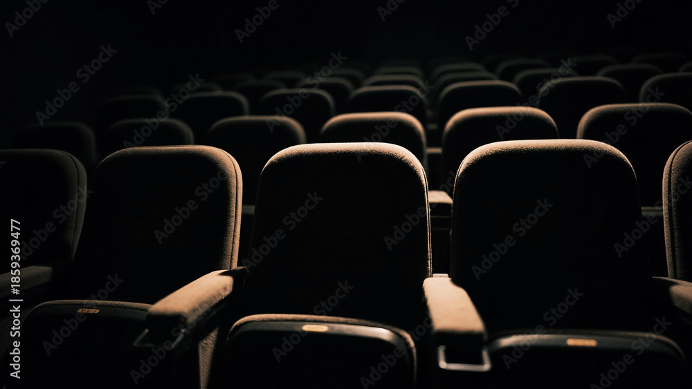 Fototapeta premium Empty rows of dark cinema seats in a movie theater, illuminated by a subtle light from above, creating a dramatic and quiet atmosphere.