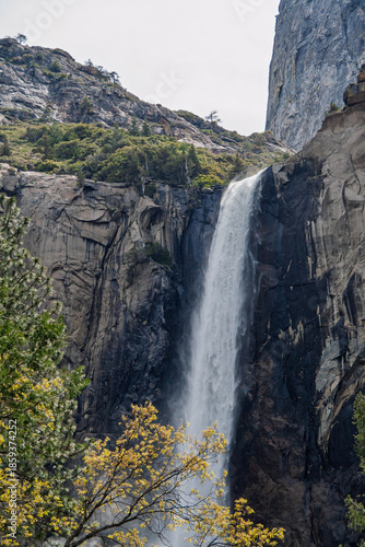 Bridalveil Fall plunges over the granite cliff face in Yosemite National Park, surrounded by a dramatic mountainous landscape with diverse tree foliage and natural rock formations