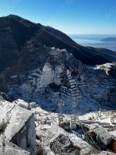 Panoramic view of white marble terraces in Carrara quarry, Italy. Steep excavation walls in Apuan Alps with Tyrrhenian coast on background. Industrial mining site, geological stone extraction, Versili