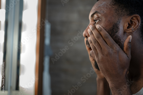 Tableau sur toile Black man looking at himself in a bathroom mirror while applying skincare around his eyes