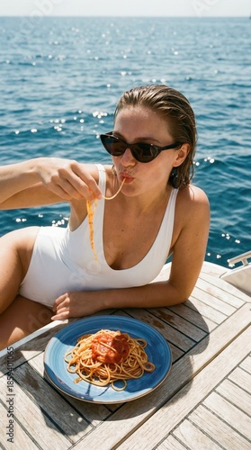 Woman eating spaghetti on yacht deck in white swimsuit with ocean background
