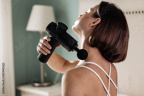 Cuadro en lienzo Woman using a vibrating massage gun on her neck in a cozy room with soft lightin