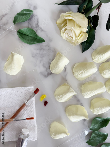 Handmade tea rose sculpted from air-dry clay lying on a table, with separate petals and green leaves nearby. Clay flower during crafting process. DIY art, handmade decoration, creative workshop